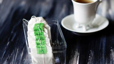 Ein Teebeutel steht in einem transparenten Behälter und wird von einer Tasse Tee umgeben.