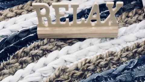 Ein gehäkelter Flickenteppich in Blau-, Weiß- und Brauntönen liegt ausgebreitet, während ein Holzschild mit der Aufschrift „RELAX“ darauf liegt.