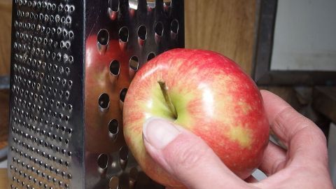 Ein roter Apfel wird mit einer Hand an einer Käsereibe gerieben, um gesunde Nahrungsmittel bei Durchfall zuzubereiten.