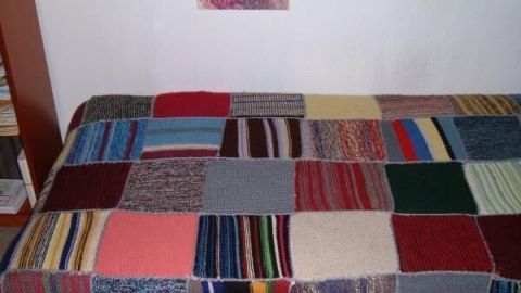 Eine gestrickte Patchworkdecke liegt ausgebreitet auf einem Bett, bestehend aus bunten Quadraten in einem Schlafzimmer.