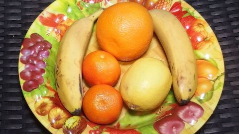 Ein Obstkörbchen mit Bananen, Orangen und einer Birne liegt auf einem bunten Teller, umgeben von einem dunklen Hintergrund.