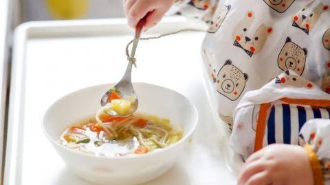 Ein kleines Kind isst mit einem an einem Löffel befestigten Faden aus einer Schüssel mit Gemüsesuppe.