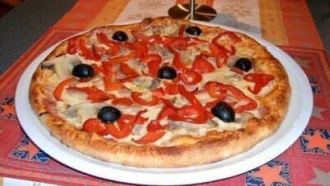 Eine frisch gebackene Pizza liegt auf einem Teller und ist mit Paprika und Oliven belegt. Der Tisch hat ein farbenfrohes Tischtuch.