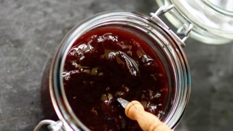 Eine Glas Marmelade mit einer tiefroten Farbe steht auf einem grauen Hintergrund. Ein kleiner Löffel steckt darin.