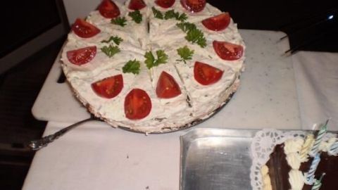 Eine Schwarzbrottorte mit Frischkäse, Tomaten und Petersilie steht auf einem weißen Tisch, daneben ein Tablett mit Kuchen.