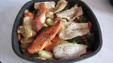 Eine Pfanne mit verschiedenen Fischfilets auf Brokkoli, Karotten und Kartoffeln, gewürzt mit Pfeffer, auf einem Tisch.