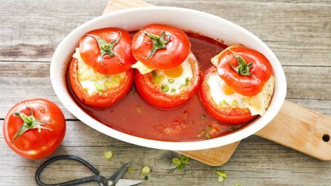 Rote Tomaten werden mit Schafskäse und Ei gefüllt, in einer Sauce gebacken und auf einem Holzbrett präsentiert.