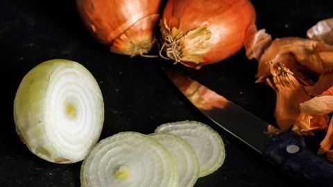 Eine geschnittene Zwiebel liegt neben ganzen Zwiebeln und einem Messer auf einer dunklen Arbeitsfl&auml;che, bereit zum R&ouml;sten.