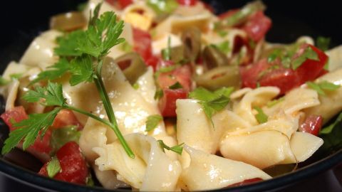 Ein bunter Nudelsalat liegt in einer schwarzen Schüssel, angereichert mit frischem Gemüse und Kräutern, leicht und lecker.
