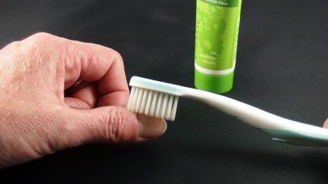 Eine Hand hält eine Zahnbürste, während sie Nagel- und Nagelbettpflege mit einer Tube in einem dunklen Umfeld durchführt.