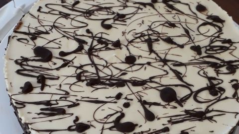 Eine Rhabarbertorte liegt auf einem weißen Servierplatte und wird von einer Holzoberfläche umgeben. Schokoladenswirbel zieren die cremige Oberfläche.