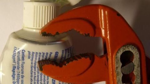 Ein orangefarbener Schraubenschlüssel drückt eine Tubenöffnung zusammen, um die Tube vollständig auszudrücken.