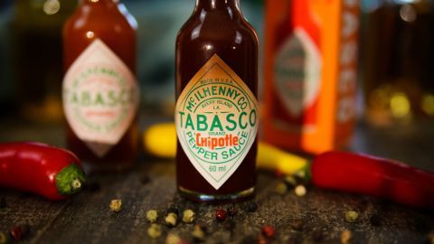 Eine Flasche Tabasco-Sauce steht im Vordergrund, umgeben von weiteren Gewürzen und chillies auf einem holzigen Tisch.