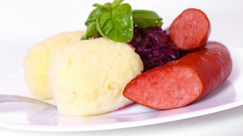 Auf einem weißen Teller liegen zwei Bratwürste neben zwei Portionen Kartoffelbrei und einem Klecks Rotkraut, garniert mit Basilikum.
