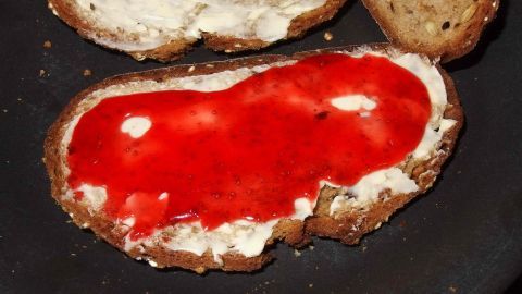 Ein Stück Brot ist mit leuchtend roter Erdbeermarmelade bedeckt, das auf einem schwarzen Teller liegt, umgeben von weiteren Brotscheiben.