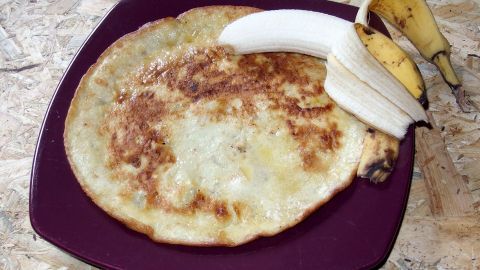 Auf einem lila Teller liegt ein goldbrauner Pfannkuchen, daneben ruhen eine ungeschälte und eine halbierte Banane, die verzehrbereit ist.