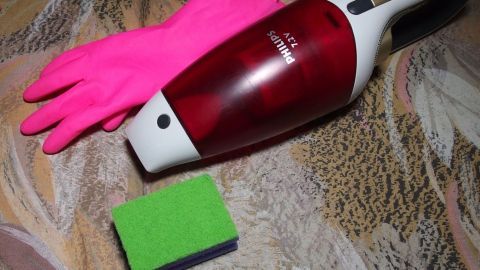 Ein roter Handstaubsauger liegt neben einem grünen Schwamm und pinken Gummihandschuhen auf einem gemusterten Untergrund.