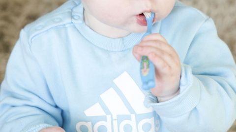 Ein kleines Kind putzt mit einer blauen Zahnbürste seine Zähne, während es in einem hellen Raum sitzt.