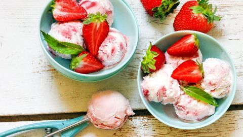 Zwei Schalen mit frischem Erdbeereis und ganzen Erdbeeren sind auf einem Holzuntergrund platziert, umgeben von einem Eisportionierer.
