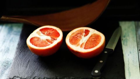 Dünnschalige Früchte sind meistens saftiger als Dickschalige. Die Dickschaligen erkennt man daran, dass sie am Stielende spitz zulaufen und rau und runzelig aussehen. Zwei halbierte Grapefruits liegen auf einem schwarzen Brett neben einem Messer, bereit zum Schneiden und genussvollen Verzehr.