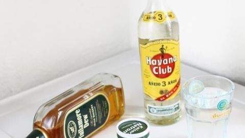 Eine Fläschchen Rum und ein Glas stehen auf einem Tisch, umgeben von einem anderen Schnaps und einer Medikamentenpackung gegen Kater.
