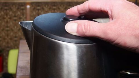 Ein Wasserkocher steht auf einer Oberfläche, während eine Hand den Deckel schließt, um Energie beim Wasserkochen zu sparen.
