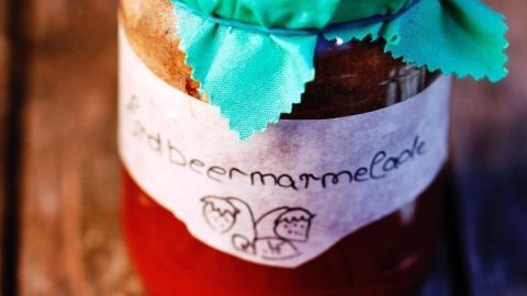 Ein Marmeladenglas mit grüner Decke und einem Etikett für "Erdbeermarmelade" steht auf einem Holztisch.