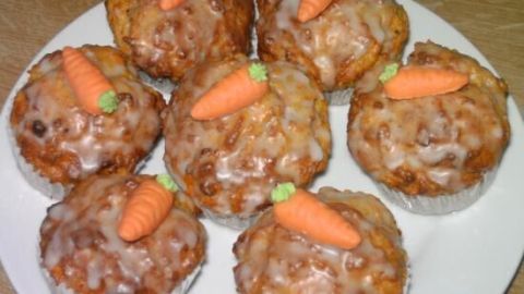 Muffins mit Zuckerguss und Marzipankarotten auf einem weißen Teller liegen auf einem Holztisch.