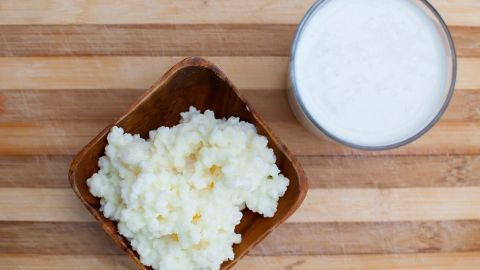 Kefirknollen liegen in einer Schüssel neben einem Glas frischem Kefir auf einer Holzoberfläche.