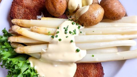 Zarte Spargelstangen liegen unter einer cremigen Soße, umgeben von Bratkartoffeln und einem Schnitzel auf einem Teller.