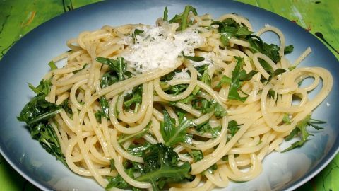 Ein Spaghetti-Rucola-Salat ist auf einem blauen Teller angerichtet, umgeben von frischem Rucola und bestreut mit Käse.
