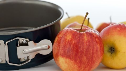 Ein rustikaler Apfelkuchen wird vorbereitet; rote und grüne Äpfel liegen neben einer Kuchenform auf einer hellen Oberfläche.