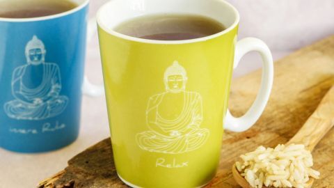 Eine grüne Tasse mit Tee steht neben einer blauen Tasse auf einem Holztablett, umgeben von Puffreis.