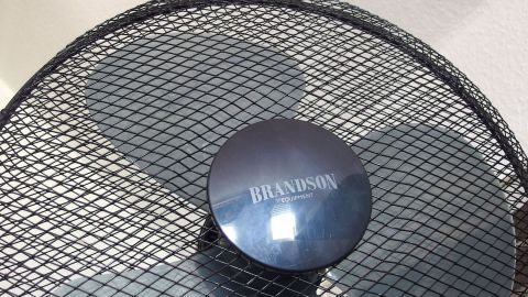 Wenn dein Ventilator unruhig läuft, dann hilft es, die Rotorblätter kurz in heißes Wasser zu tunken, wodurch sich das Plastik entspannt. Ein Ventilator mit einem runden Kopf steht still da, umgeben von einem metallenen Schutzgitter in einem neutralen Raum.