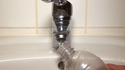 Eine transparente Trinkflasche wird unter einem Wasserhahn gehalten, der Wasser ins Bad fließt, um sie zu füllen.