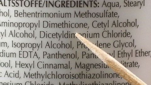 Eine Hand hält einen Holzstab, der auf eine Liste von Inhaltsstoffen auf einer Haarpflegeverpackung zeigt, um Trockenshampoo-Alternativen zu entdecken.