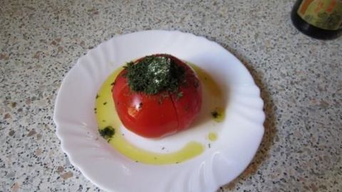 Eine rote Tomate liegt auf einem weißen Teller, garniert mit einer Dillhaube, umgeben von einem Ölring.