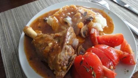 Eine saftig geschmorte Lammhaxe liegt auf einem Teller, umgeben von Püree und frischen Tomaten, perfekt angerichtet.