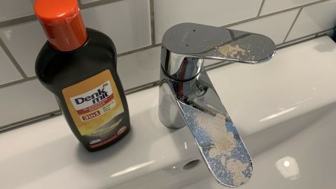 Haarfarbe auf die Badezimmerarmaturen bekommen? Mithilfe von Glaskeramikfeldreiniger bekommst du die Farbe sehr gut wieder entfernt. Ein glänzender Badezimmerarmatur mit Farbspuren, daneben eine Flasche Reiniger, zeigt den Prozess zum Entfernen von Haarfarbe.