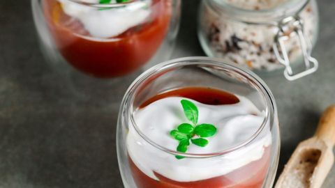 Eiskalte Gläser mit Tomatencappuccino stehen auf einem Tisch, garniert mit Sahne und einem Basilikumblatt.