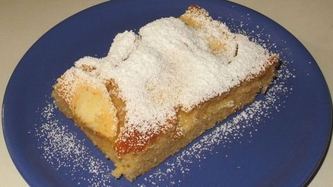 Ein leckerer Apfelkuchen liegt auf einem blauen Teller, bestäubt mit Puderzucker und umgeben von einer einfachen, hellen Oberfläche.