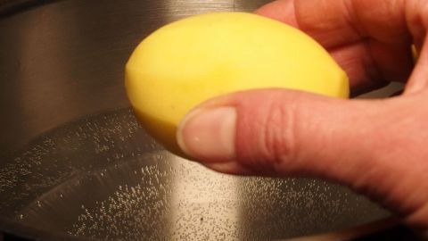 Eine Hand hält eine Kartoffel über einem Topf mit heißem Wasser, bereit zum Kochen. Dampf steigt auf.