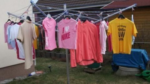 Eine Wäschespinne mit bunten T-Shirts hängt im Freien, während die Sonne scheint und eine grüne Wiese im Hintergrund sichtbar ist.