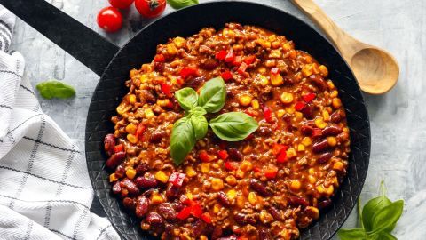 Ein schwarzer Topf enthält Chili con Carne mit roten Bohnen, frischen Tomaten und Basilikum, umgeben von einem Tischset.
