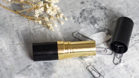 Leere Lippenstifthüllen liegen auf einer grauen Oberfläche, umgeben von Büroklammern und etwas trockenen Blumen.