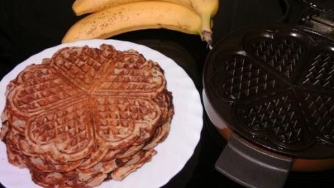 Bananen liegen neben einem Teller mit herzförmigen Waffeln auf einem Tisch, begleitet von einem offenen Waffeleisen.