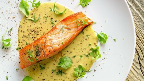 Ein Stück Lachs liegt in einer cremigen Senfsoße auf einem weißen Teller, umgeben von frischen Kräutern und Gewürzen.