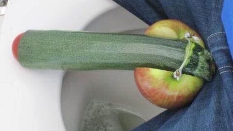 Eine Zucchini liegt auf einem Apfel in der Nähe einer Toilettenöffnung, umgeben von Jeansstoff.