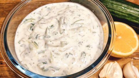 Eine Schüssel mit frischem Tzatziki steht im Vordergrund, umgeben von einem Stück Zitrone, Knoblauch und einer Gurke.