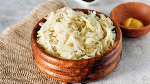 In einer Holzschüssel liegt frisch zubereiteter Weißkrautsalat auf einem neutralen Untergrund, umgeben von Gewürzen.